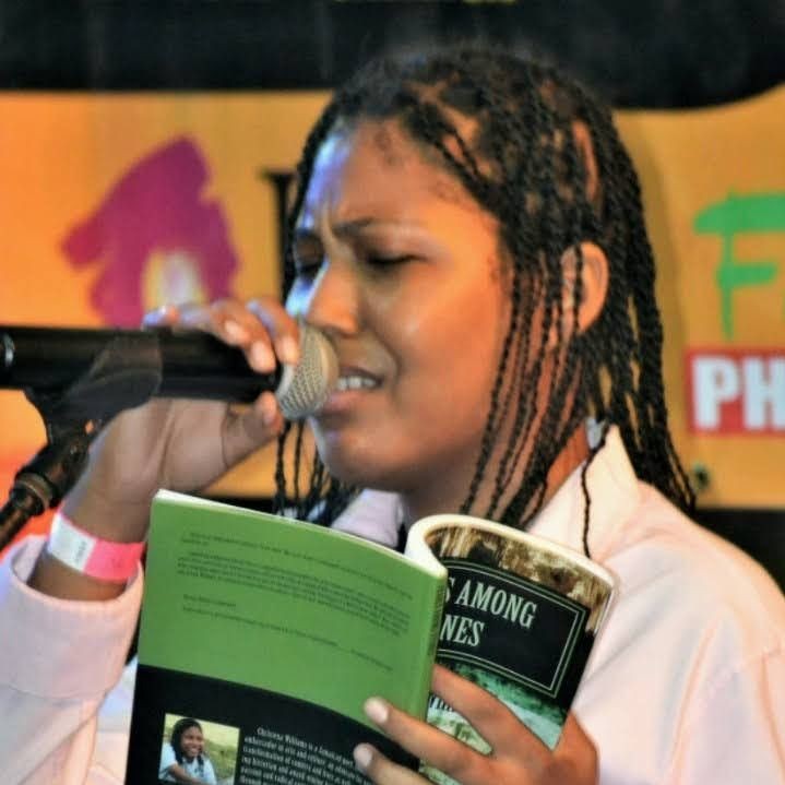 Person with braided hair holding a book and singing into a microphone on stage.